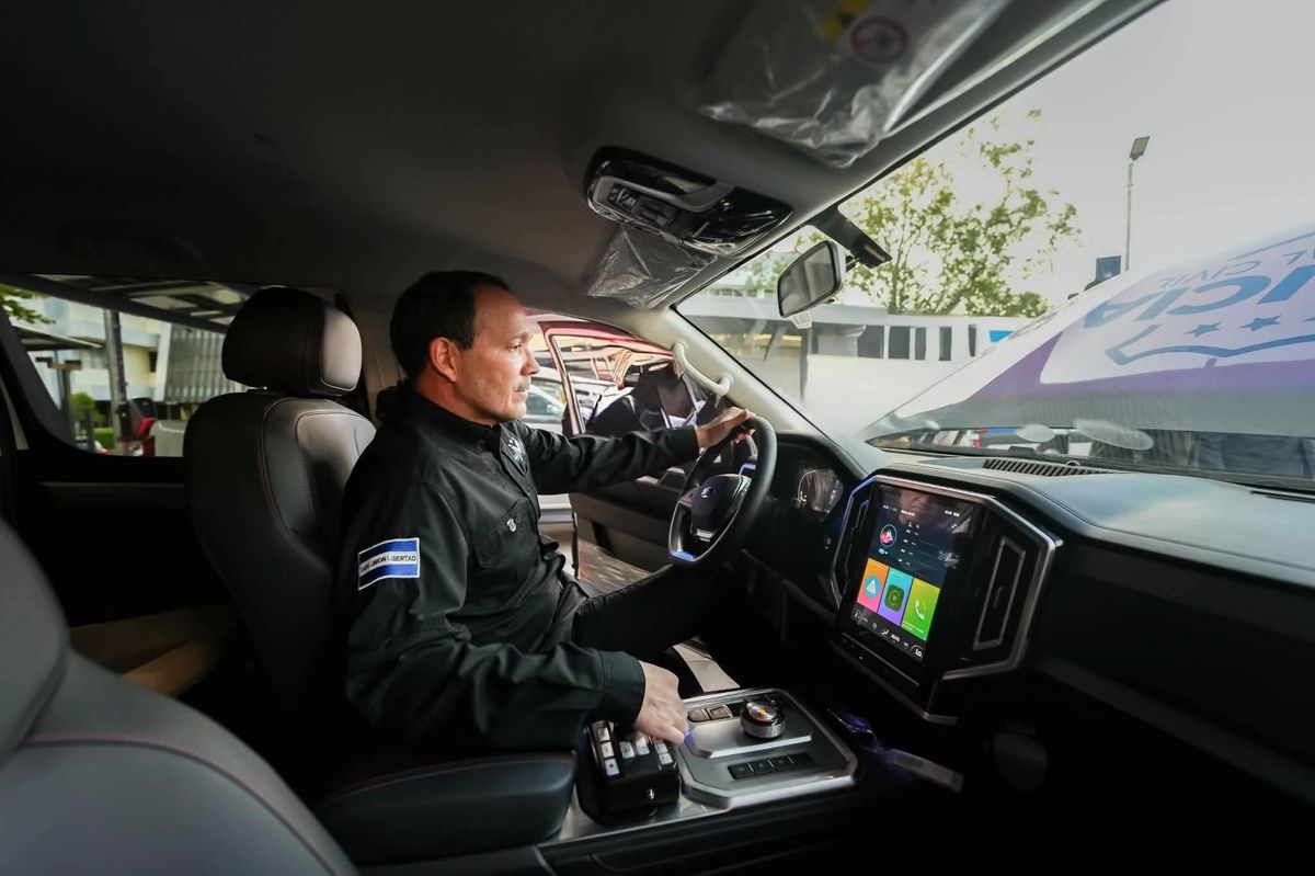 Gustavo Villatoro, vestido de negro con un parche de la bandera de El Salvador en el brazo, sentado al volante de una pickup policial nueva. Con una mano sostiene el guía y con la otra opera los controles de la consola central, junto a una pantalla táctil grande en el tablero. El parabrisas conserva el rótulo invertido de Policía Nacional Civil y el interior mantiene los plásticos protectores de fábrica.