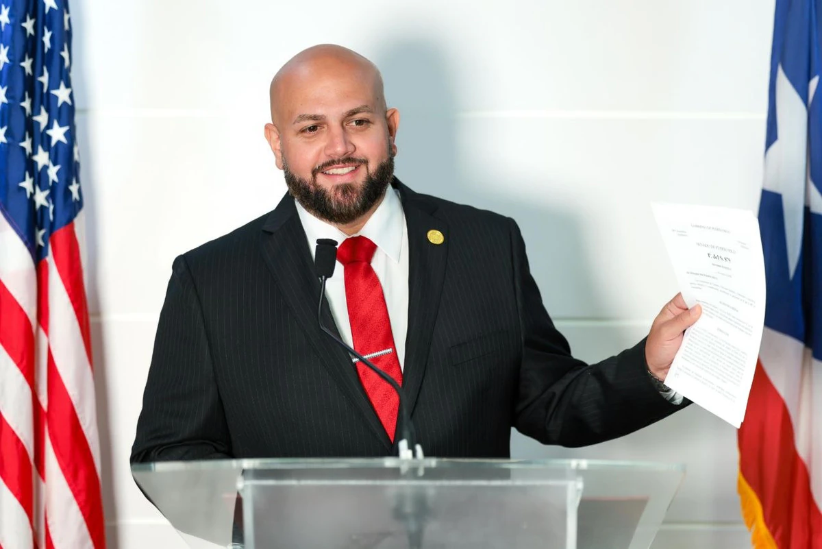 Jeison Rosa Ramos, senador del Distrito de Mayagüez-Aguadilla, de pie ante un podio transparente con micrófono, sonriente, calvo y con barba, vestido con traje negro a rayas, camisa blanca y corbata roja, con un pin dorado en la solapa, sosteniendo en alto con la mano derecha un documento con el membrete del Senado de Puerto Rico, frente a una pared blanca con la bandera de los Estados Unidos a la izquierda y la de Puerto Rico a la derecha.