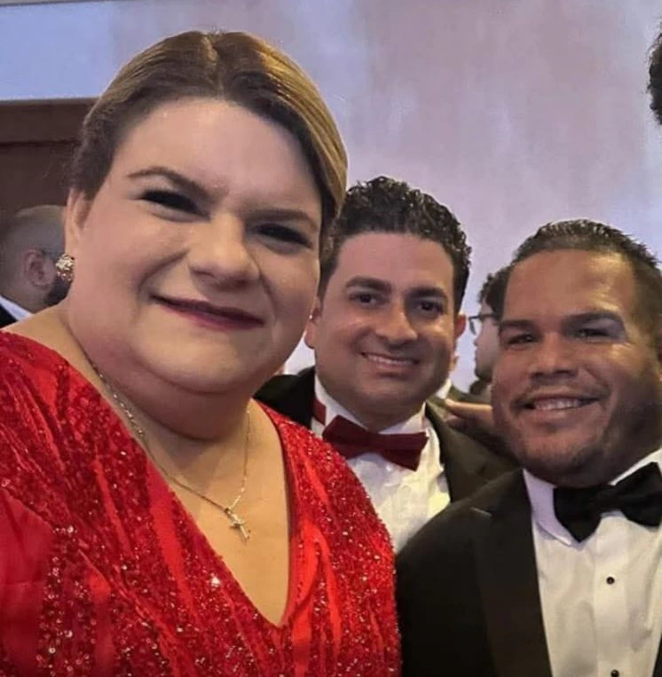 Selfie en un salón de gala. A la izquierda, la gobernadora Jenniffer González Colón, con el cabello recogido, vestido rojo de lentejuelas y una cruz al cuello, sonríe a la cámara. En el centro, José Yovín Vargas, de esmoquin con lazo rojo. A la derecha, Félix Plaud De Choudens, de barba corta y esmoquin con lazo negro. Al fondo, otros invitados y el techo iluminado del salón.