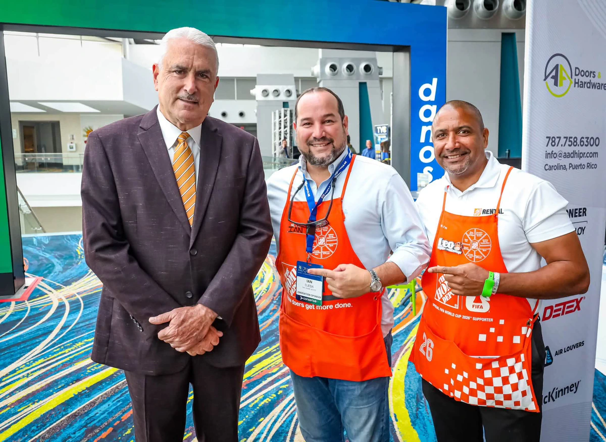 Un hombre de traje marrón y corbata amarilla a rayas posa junto a dos hombres con delantales anaranjados de The Home Depot en el vestíbulo de un centro de convenciones, sobre una alfombra de colores y con un rótulo de AA Doors & Hardware al fondo.