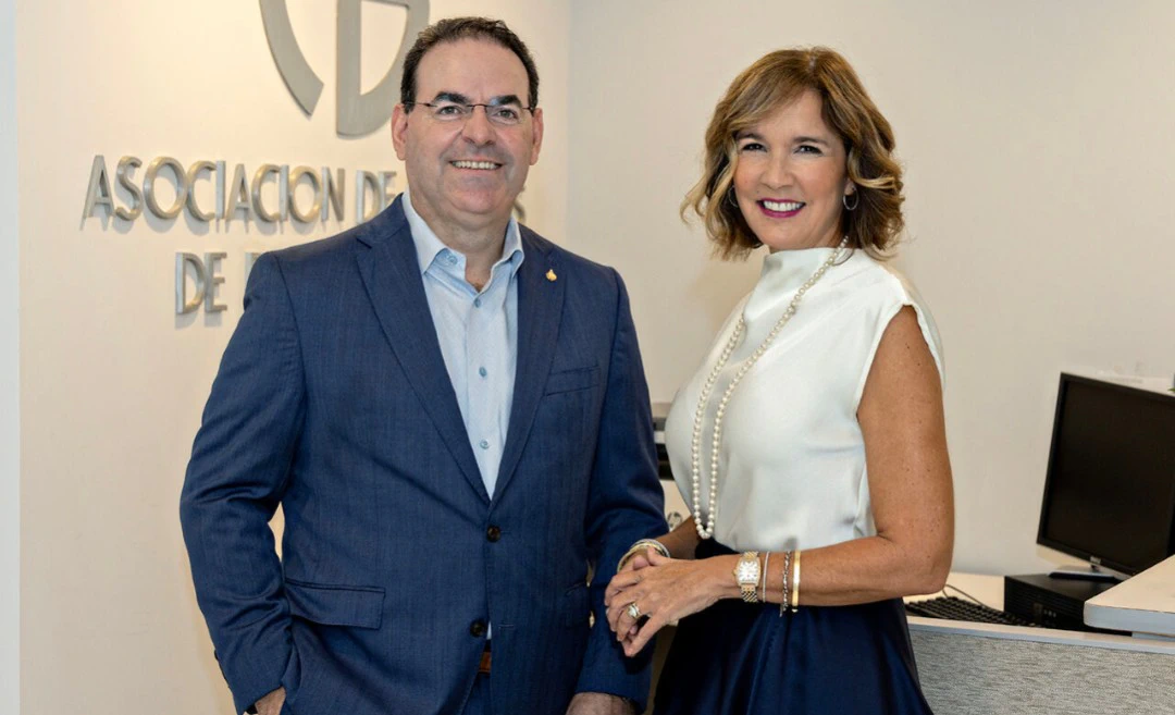 Jorge J. García, un hombre de cabello oscuro con espejuelos, traje azul y camisa celeste sin corbata, y Zoimé Álvarez Rubio, una mujer de cabello castaño con blusa blanca sin mangas, falda azul marino y collar de perlas, posan sonrientes frente a un letrero plateado con el logo y el nombre de la Asociación de Bancos de Puerto Rico.