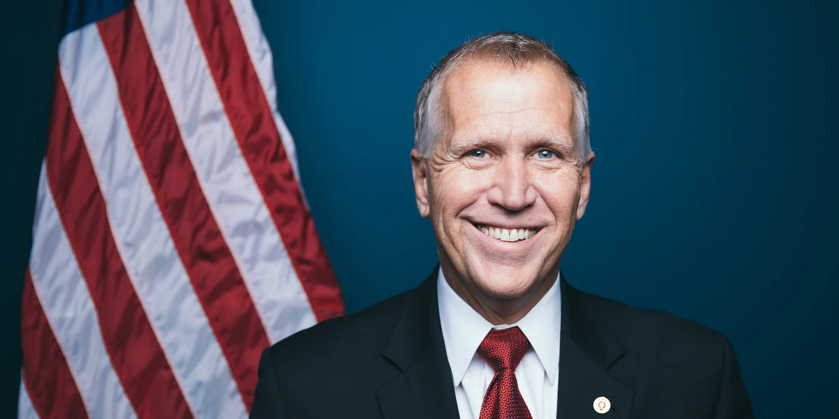 Retrato oficial del senador Thom Tillis, un hombre de cabello canoso que sonríe a la cámara, con traje oscuro, camisa blanca y corbata roja con patrón y un pin en la solapa, frente a un fondo azul con la bandera de Estados Unidos a la izquierda.